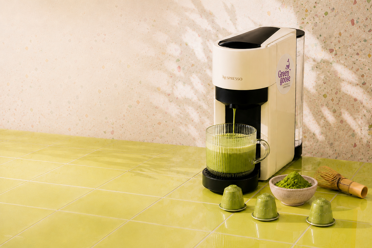 A Nespresso coffee machine dispensing green matcha tea into a glass cup, set on a glossy yellow-green tiled countertop against a speckled beige wall. Three green coffee pods, a small bowl of matcha powder, and a bamboo whisk are arranged nearby.
